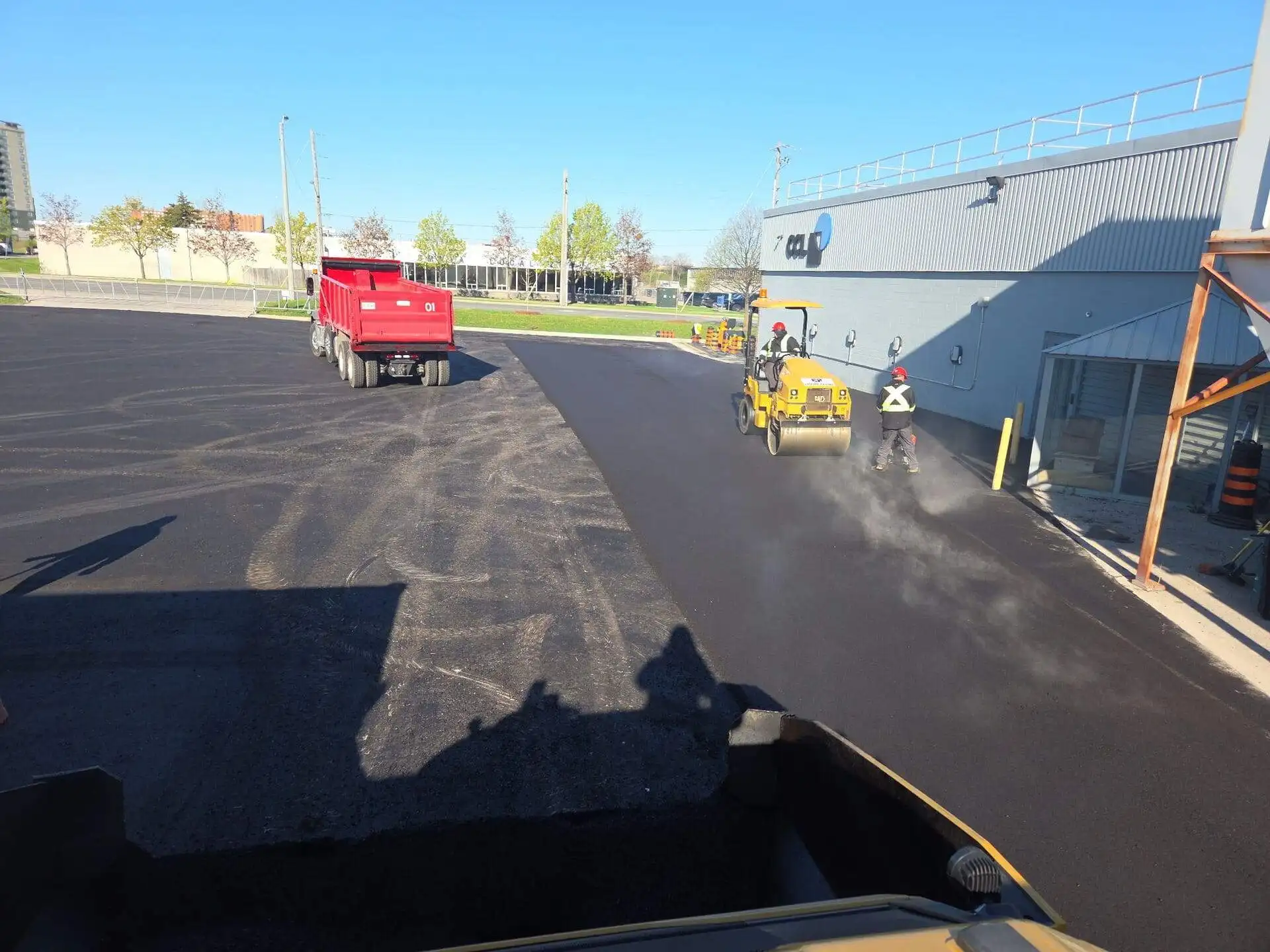 Fresh asphalt being compacted by a roller near a commercial building while workers complete finishing touches and a red dump truck is positioned nearby on a newly paved surface.