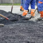 Close-up of workers using shovels and rakes to spread hot asphalt during a roadway repair project.