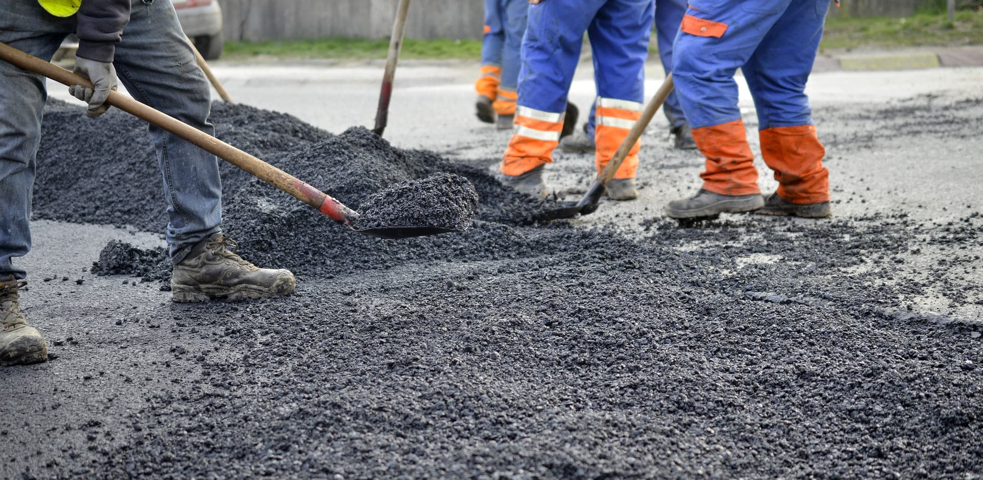 Close-up of workers using shovels and rakes to spread hot asphalt during a roadway repair project.