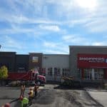 Commercial parking lot resurfacing in progress at a retail plaza, featuring asphalt trucks, paving equipment, and crew working in front of storefronts.