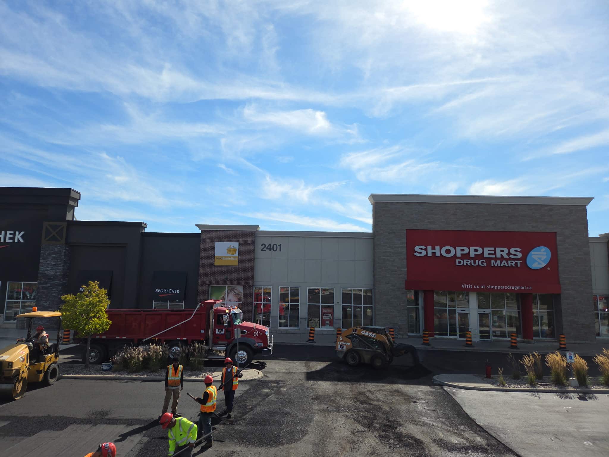 Retail Asphalt Paving Commercial parking lot resurfacing in progress at a retail plaza, featuring asphalt trucks, paving equipment, and crew working in front of storefronts.