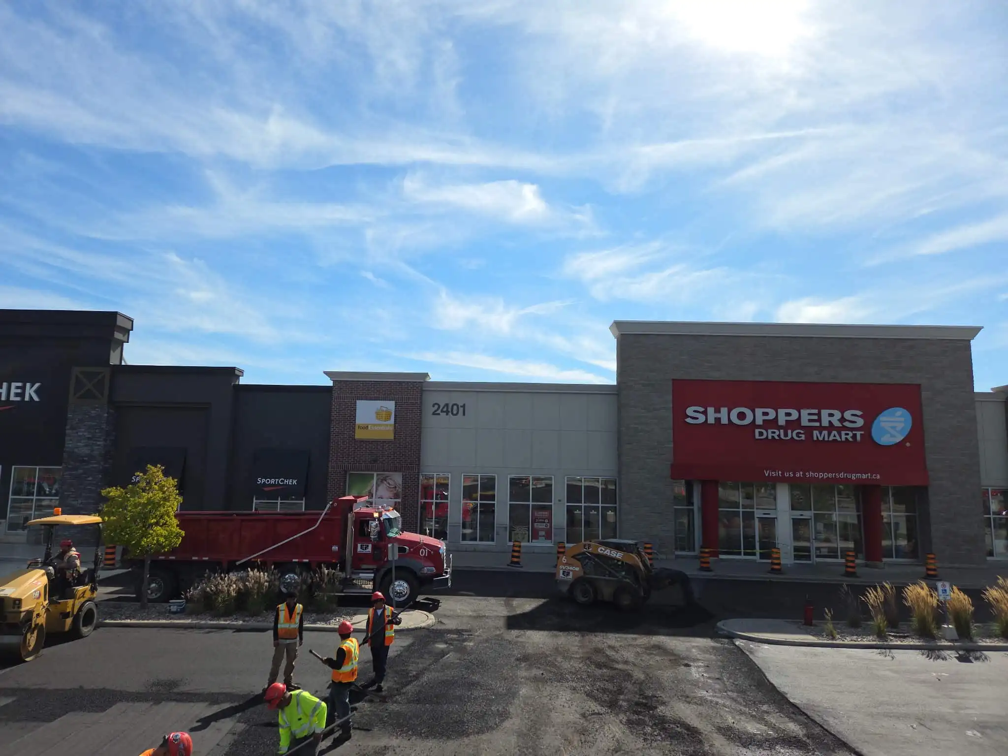 Commercial parking lot resurfacing in progress at a retail plaza, featuring asphalt trucks, paving equipment, and crew working in front of storefronts.