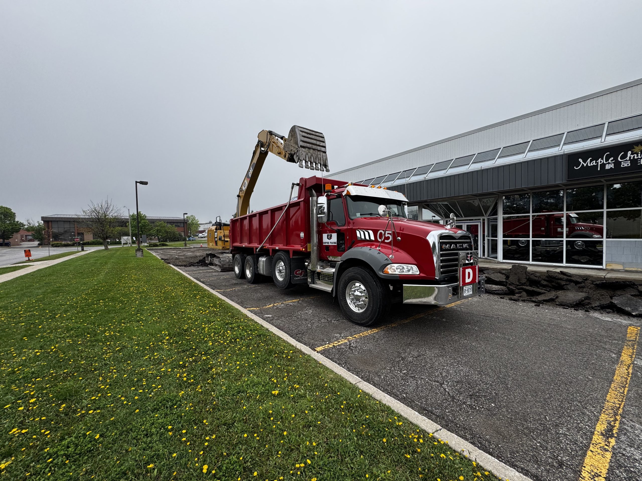 Prepare Asphalt Budgets After Winter Empire Paving red dump truck parked in a commercial parking lot in Ontario while an excavator loads removed asphalt into the truck during parking lot reconstruction.