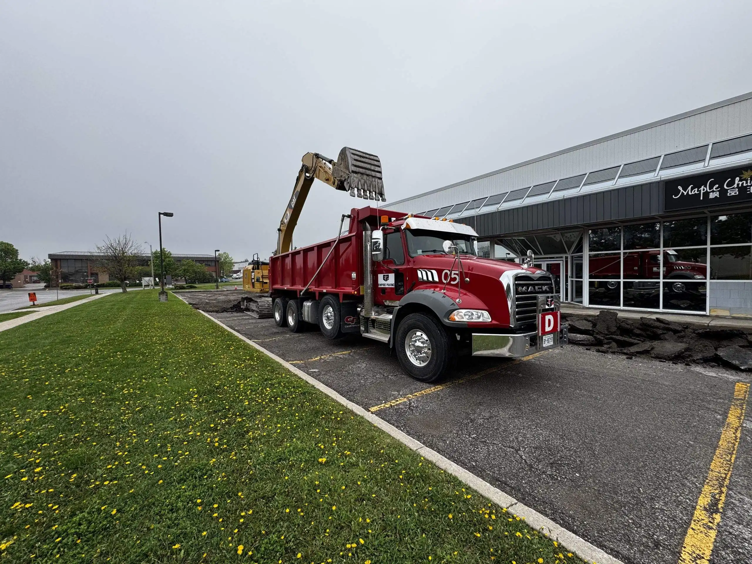 Prepare Asphalt Budgets After Winter Empire Paving red dump truck parked in a commercial parking lot in Ontario while an excavator loads removed asphalt into the truck during parking lot reconstruction.