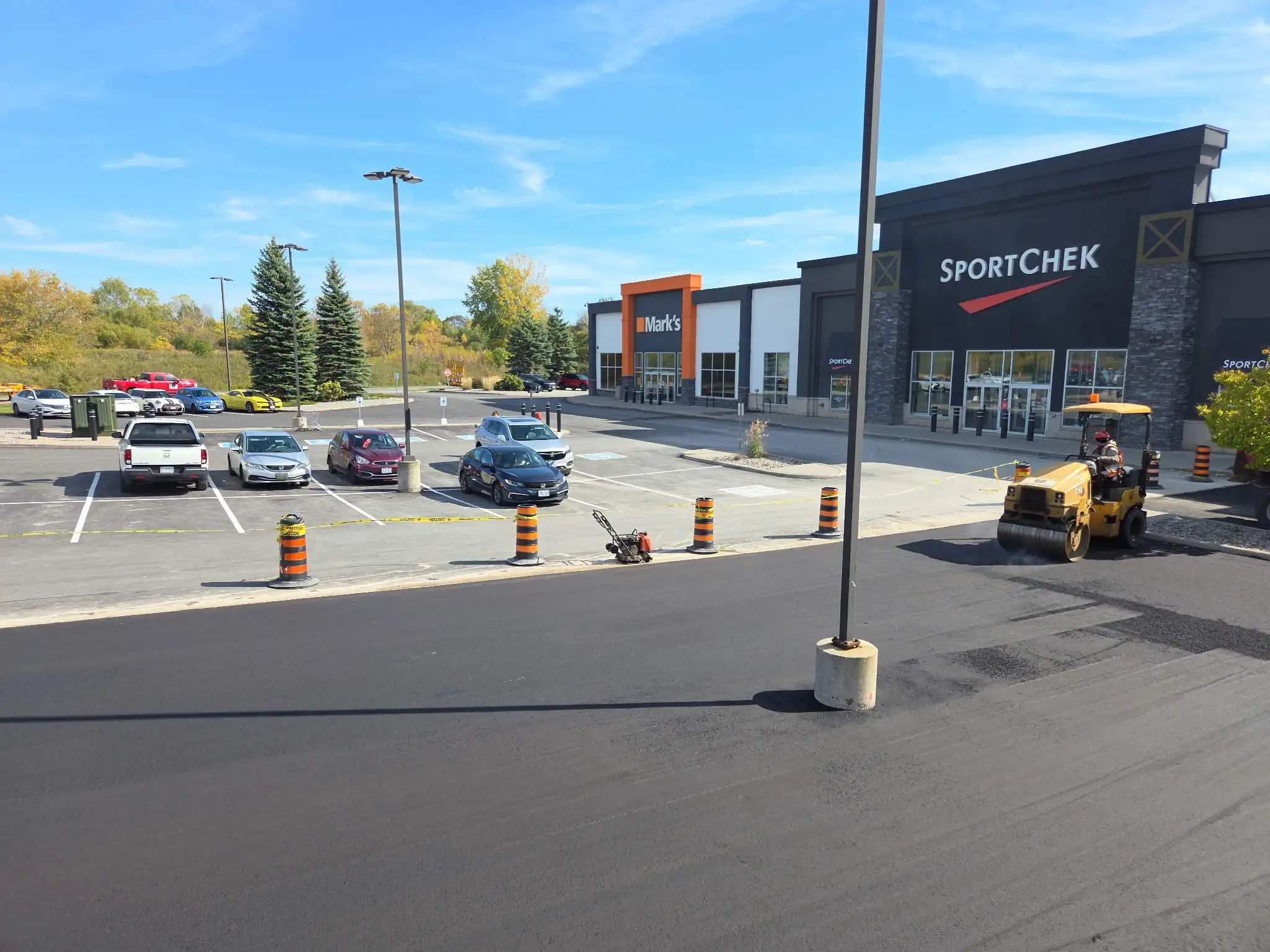 Fresh asphalt paving in progress at a commercial shopping plaza, with a roller compacting the surface near retail stores and a newly paved parking lot.