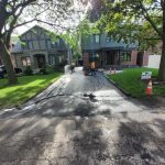 Residential driveway undergoing asphalt maintenance with workers applying sealcoating and preparing the surface in a suburban neighborhood.
