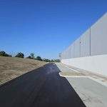 Large-scale asphalt paving project alongside a commercial building, with a newly laid smooth roadway being compacted by rollers under clear blue skies.