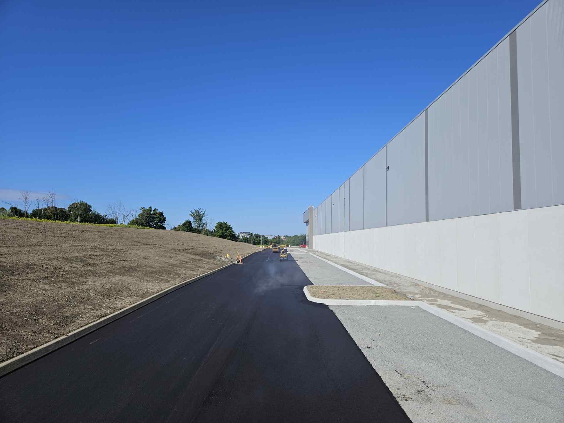 Large-Scale Asphalt Projects Large-scale asphalt paving project alongside a commercial building, with a newly laid smooth roadway being compacted by rollers under clear blue skies.