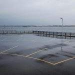 Wet commercial asphalt parking lot along a waterfront railing showing water accumulation from trapped organic debris.