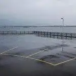 Wet commercial asphalt parking lot along a waterfront railing showing water accumulation from trapped organic debris.