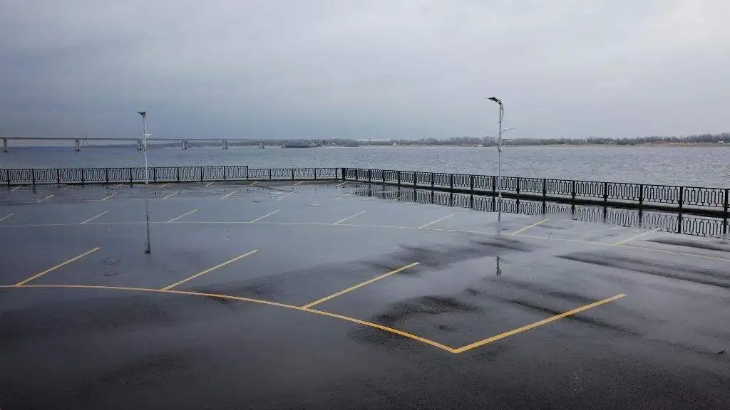 Wet commercial asphalt parking lot along a waterfront railing showing water accumulation from trapped organic debris.
